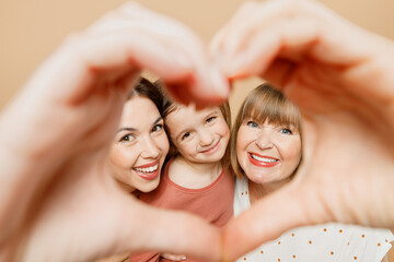 Obraz na płótnie Canvas Happy women wearing casual clothes with child kid girl 6-7 years old. Granny mother daughter show shape heart look through hand heart-shape sign isolated on plain beige background. Family day concept.