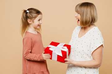Side view women wearing casual clothes with child kid girl 6-7 years old. Grandmother granddaughter hold present box with gift ribbon bow isolated on plain beige background Family parent day concept.