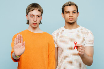 Young couple two gay men wear casual clothes together hold AIDS awareness red ribbon show stop palm gesture isolated on plain blue background studio portrait. Pride day june month love LGBTQ concept.
