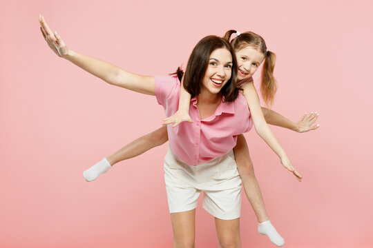 Happy woman wearing casual clothes with child kid girl 6-7 years old. Mother, give piggyback ride to joyful daughter sitting on back isolated on plain pastel pink background Family parent day concept