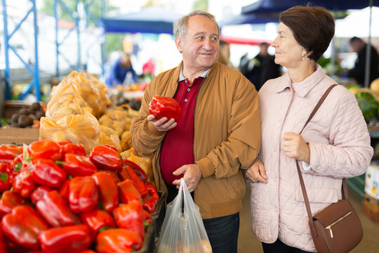 Positive senior couple in casual wear picking bell peppers during shopping at crowded bazaar