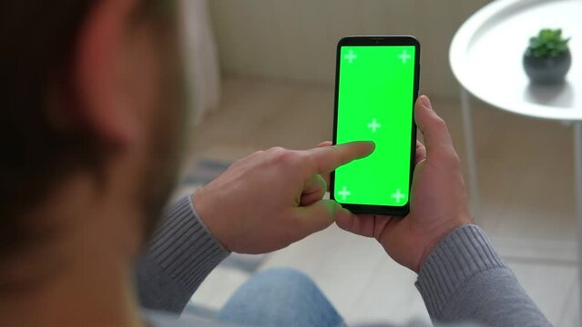 Young man holding smartphone green mock-up screen in hand. Male person using chroma key mobile phone. Vertical mode. Touching, swiping display, tapping, surfing Internet social media. Gesturing