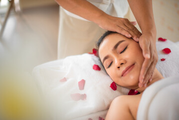 Fototapeta na wymiar Beautiful asian woman is receiving a head massage from a highly skilled professional masseuse on a bed adorned with sprinkled roses in a spa room.