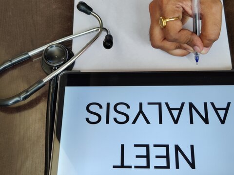 Picture of a person writing on a paper with a pen with NEET ANALYSIS written on a screen. NEET is a medical entrance exam in India in which many candidates appear every year.