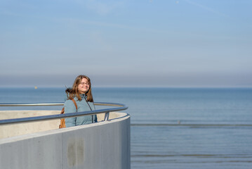Smiling young woman in blue parka walking near sea at sunny spring day. Enjoying moment
