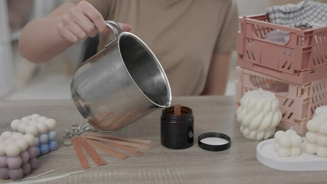 woman making candles at home, close up of female hands pouring yellow liquid wax from pan into glass bottle form