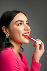 alluring woman with trendy earrings and shiny brunette hair applying red lipstick and smiling while looking away and and preparing for party isolated on grey background