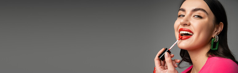 alluring woman with trendy earrings and brunette hair applying red lip gloss and smiling while looking at camera and posing isolated on grey background, banner