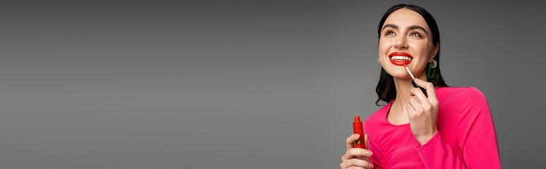 stunning young woman with brunette hair and trendy earrings posing in magenta party dress while applying red lip gloss and smiling isolated on grey background, banner