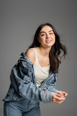 happy young woman with brunette hair and flawless natural makeup standing in fashionable denim jacket while smiling while posing isolated on grey background