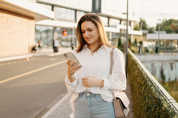 Young woman wearing white shirt is looking at the phone while waiting for a taxi.