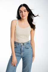 Fototapeta premium gorgeous woman with brunette hair, perfect skin and natural makeup standing in denim jeans and tank top while looking at camera on white background