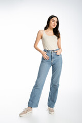 Obraz premium full length of enchanting woman with natural beauty, brunette hair and perfect skin smiling while posing with hand in pocket while standing in denim jeans and tank top on white background