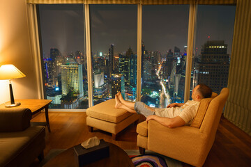 Man resting on chair in cozy calm room interior in the evening and looks through window and enjoy of modern Bangkok metropolis city lights and beautiful urban panoramic view at night from the high - obrazy, fototapety, plakaty