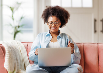 beautiful young smiling ethnic woman with curly hair uses laptop smiling, gesticulating and talking to online while sitting on sofa