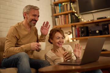 Happy mature couple waving to someone during video call at home
