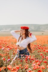 Happy woman in a poppy field in a white shirt and denim skirt with a wreath of poppies on her head posing and enjoying the poppy field.