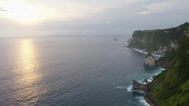 Aerial Sunset view Tropical seaside of Nusa Penida Island high steep coastal cape edges and Coves overlooking the waves of  indian ocean crashing down the green vegetated cliff, Indonesia.