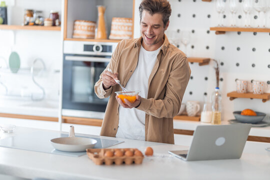 A smiling male freelancer prepares eggs for an omelet and works on the laptop.