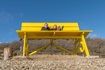 A middle aged couple is sitting on a giant yellow bench surrounded by nature