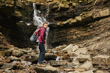 Obraz premium Hiking in mountains. Woman enjoying hike on sunny vacation day. Female with backpack walking close to waterfall. Spending summer vacation close to nature