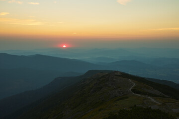 Fototapeta premium Sunrise in mountains. Natural mountain landscape with illuminated misty peaks, foggy slopes and valleys, blue sky with orange yellow sunlight