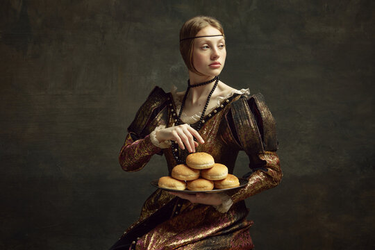 Portrait of young, elegant, beautiful girl in vintage costume, dress holding burger buns against dark green background. Concept of history, renaissance art, comparison of eras, health and food