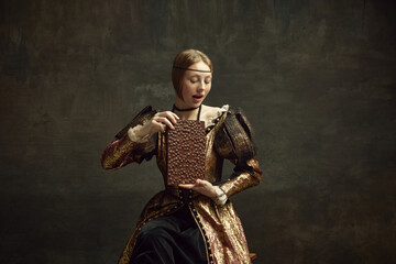 Fototapeta premium Portrait of young girl, medieval royal person, pretty princess in vintage dress holding giant chocolate bar over dark green background. Concept of history, renaissance art remake, comparison of eras