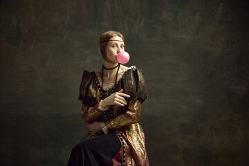 Portrait of pretty young girl, medieval princess in vintage dress posing with bubble gum against dark green background. Concept of history, renaissance art remake, comparison of eras, modernity - obrazy, fototapety, plakaty