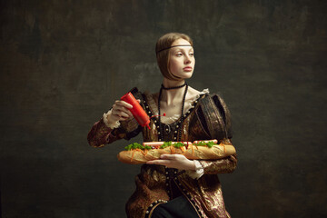 Portrait of young girl, queen, princess in vintage costume putting ketchup on giant sandwich baguette on dark green background. Concept of history, renaissance art, comparison of eras, health and food