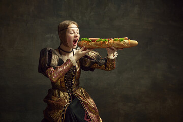 Portrait of young girl, princess in vintage costume eating giant baguette sandwich against dark green background. Concept of history, renaissance art, comparison of eras, health and food, diet