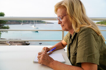 a left-handed old woman writes concentrated in the notebook in seascape. international left handers day