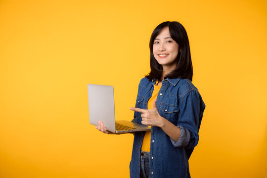 portrait young happy woman wearing yellow t-shirt and denim shirt holding laptop and point finger to screen isolated on yellow studio background. business technology application communication concept.