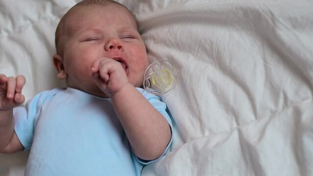 Cute newborn baby with pacifier lying in cradle at home, top view. Charming infant suckling pacifier nipple and spit it out. Child one month old lies in bed on white blanket. Copy Space. Blue bodysuit