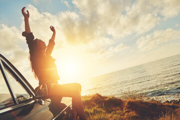 Road trip, sunset and arms raised with a woman at the coast, sitting on her car bonnet during...