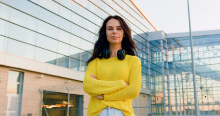 Front view of gorgeous smiling stylish young woman in trendy clothes which looking into camera with crossed arms near big urban modern building