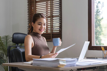 Happy Asian businesswoman working and checking work with laptop computer while having a comfortable cup of coffee at office.