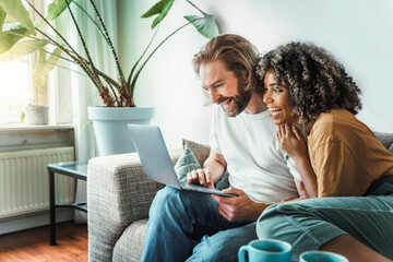 Multiracial young couple watching computer laptop sitting on the sofa at home - Happy diverse husband and wife using pc online services - Technology life style concept - 601355561