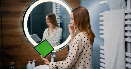 Happy woman brushing teeth and looking on tablet green screen pc in modern bathroom. Beautiful girl...