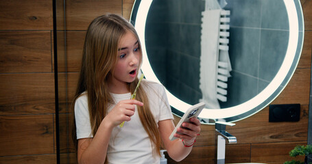 Cute little girl brushing teeth and using smartphone checking social networks in bathroom. Girl having mobile phone in hand and writes a text message.