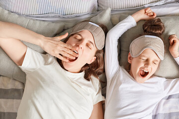 Sleepy mother and daughter sleeping together waking up wearing sleep masks and white shirts yawning...
