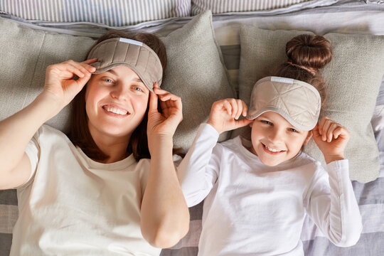 Mother and daughter enjoyed lazy morning together in cozy bedroom, wake up slowly, taking off blindfolds looking at camera smiling expressing positive emotions.