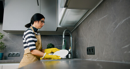 Serious young woman in yellow gloves washing white plate dishes at the modern kitchen at home
