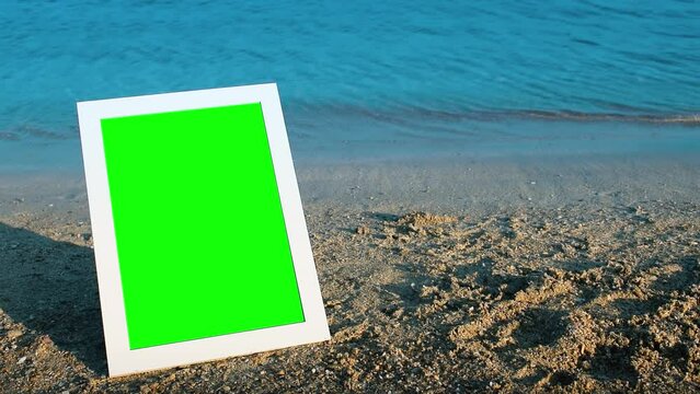 White photo frame standing on the beach by the sea. Green screen.