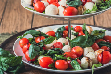 Classic italian salad Insalata Caprese with cherry tomatoes, mini mozarella pearls, basil leaves and balsamic glaze served on a cake stand, close up