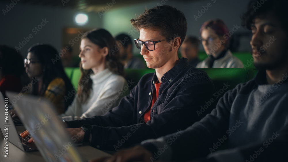 Poster Young Male Student Studying in University with Diverse Multiethnic Classmates. He is in Uplifting Mood and is Using a Laptop Computer. Patiently Listening to Professor's Lecture and Taking Notes