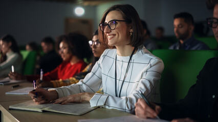 Thoughtful Beautiful Young Female Studying in University, Listening to a Lecture in a Crowded Room with Diverse Multiethnic Classmates. Students Listening to Lecture and Taking Notes in Notebook