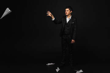 Young mafia member with a slicked back hair, dressed in black suit hold and throws a paper plane. Italian mafia gangster. isolated on black background.