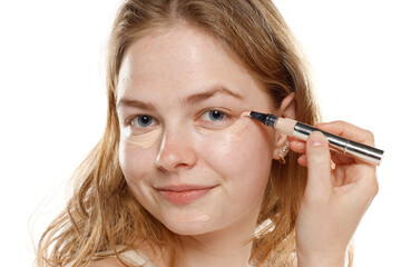 young blond woman apply concealer under the eye on a white studio background