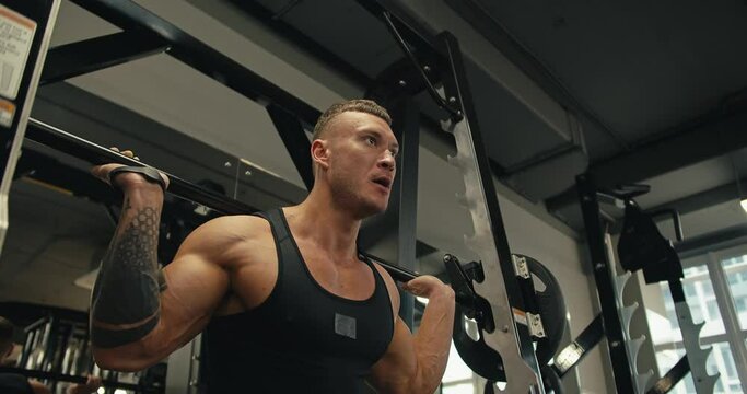 A young athlete in a black jersey squats with a barbell on a special machine to improve leg muscles. Leg day at the gym, exercises to strengthen the muscles of the legs